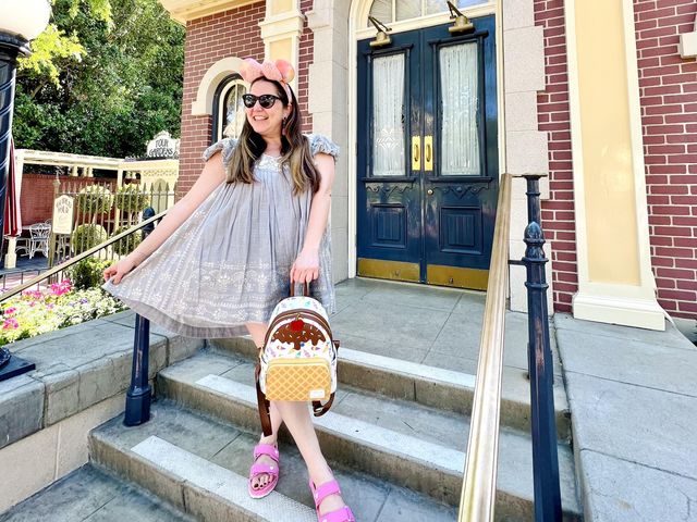 A woman stands on a stoop, holding out her skirt in one hand and an ice cream-themed mini backpack in the other, while smiling and wearing sunglasses.
