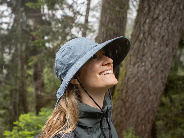 A woman wears a wide-brimmed rain hat in a forest while looking up and smiling.