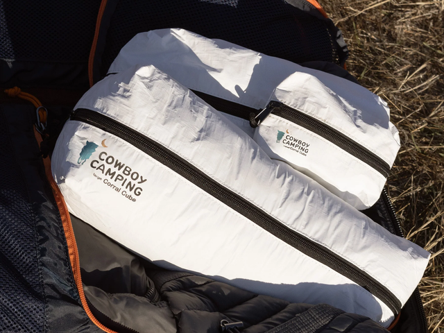 Two white rugged packing pouches sitting against a bag and grass.