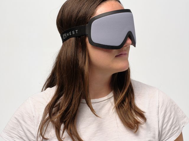 A woman wears a goggle-shaped eye mask while standing against a white background.
