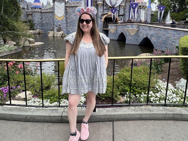 A woman wearing Minnie ears and sunglasses poses in front of the castle at Disneyland and smiles for the camera, pointing out her feet wearing pink sneakers.