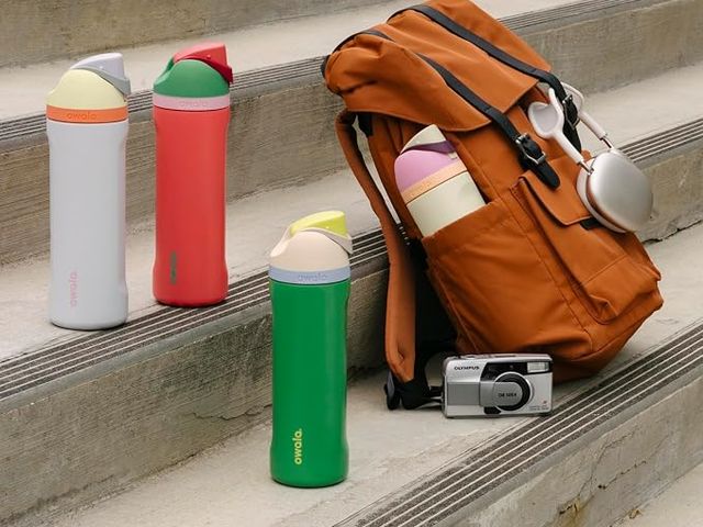 A backpack on concrete steps with a few colorful Owala water bottles around it and a camera.