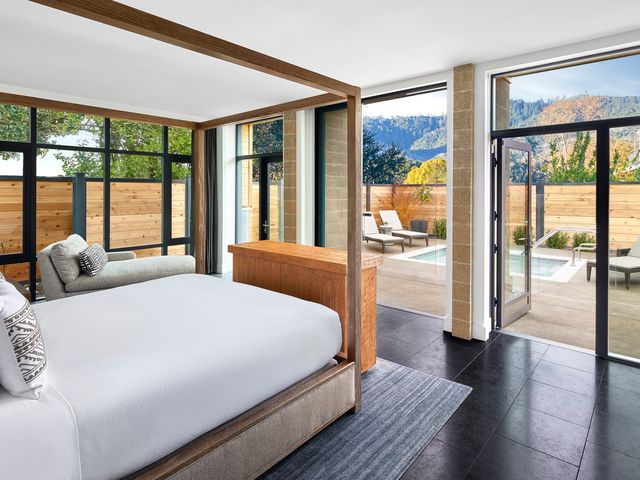 A modern looking hotel room where a bedroom opens up to a terrace with a pool lined with chairs and hilly views. 