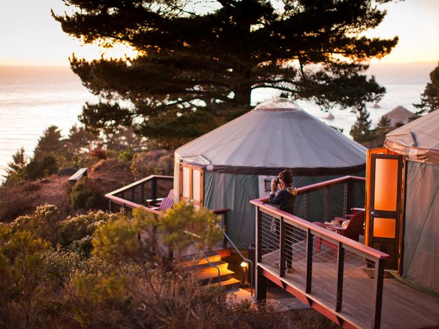 Two yurt tents amid trees facing the Pacific Ocean with a woman on one deck looking at the view through binocular.