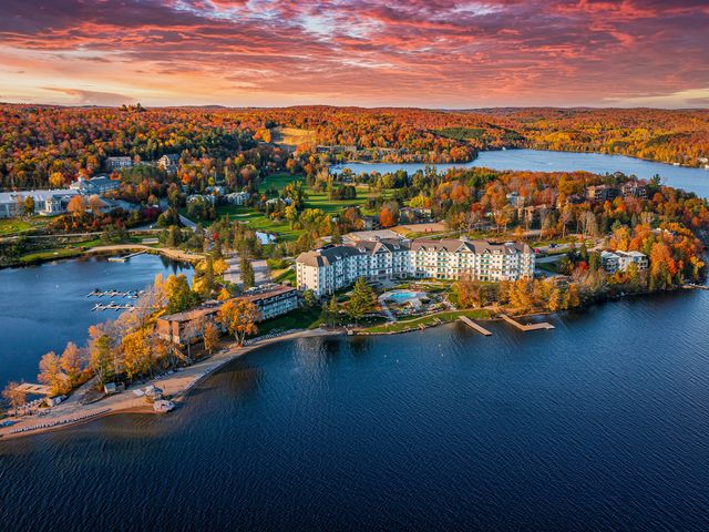A stunning fall foliage aerial of Muskoka, with buildings, plenty of nature, and the beauty of the clear blue water.