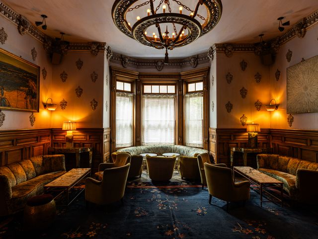 A dimly lit room with a bay window, antique upholstered sofas, velvet chairs, an ornate rug, and a chandelier.