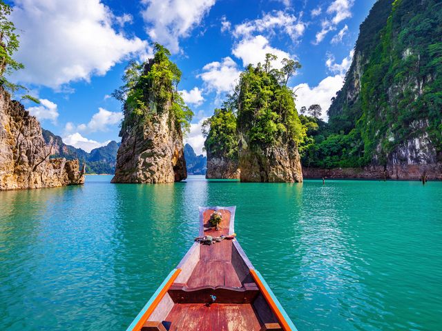 A view of rock pillars and mountains from a boat atop blue water in Khao Sok National Park in Thailand.