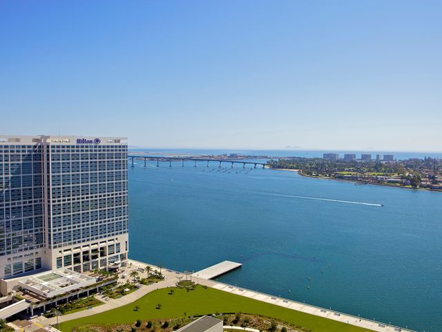 The large building exterior of the Hilton San Diego Bayfront next to a green grassy park and facing a harbor with blue water.