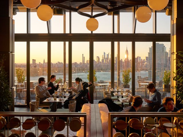 A contemporary bar lounge area with pendant lamps and many people sitting at small tables next to floor to ceiling windows with views of Manhattan with light streaming in at sunset.