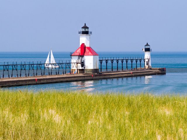 A long pier juts out into the water with two lighthouse structures on it, as seen from a grassy shore.