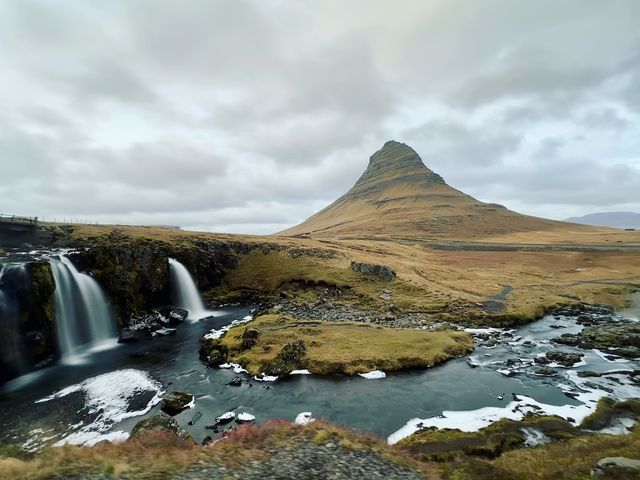 A river in the foreground with two small waterfalls flowing into it, with green mossy fields behind it and a mountain rising above.