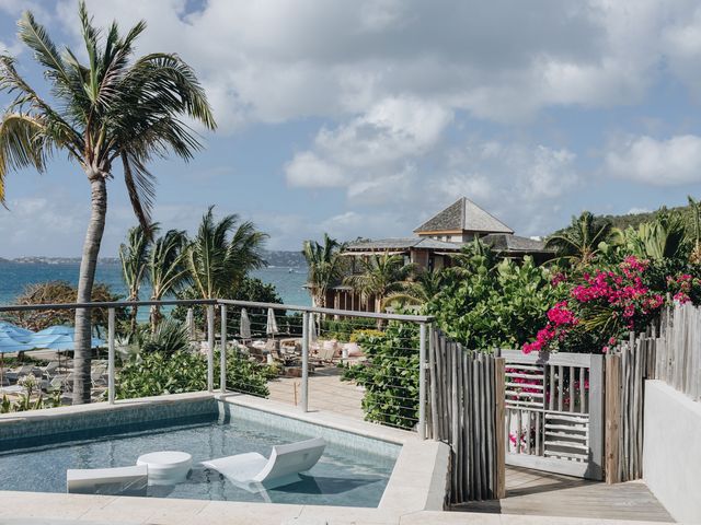 A small pool on a deck facing a beach area with loungers and umbrellas framed by palm trees.