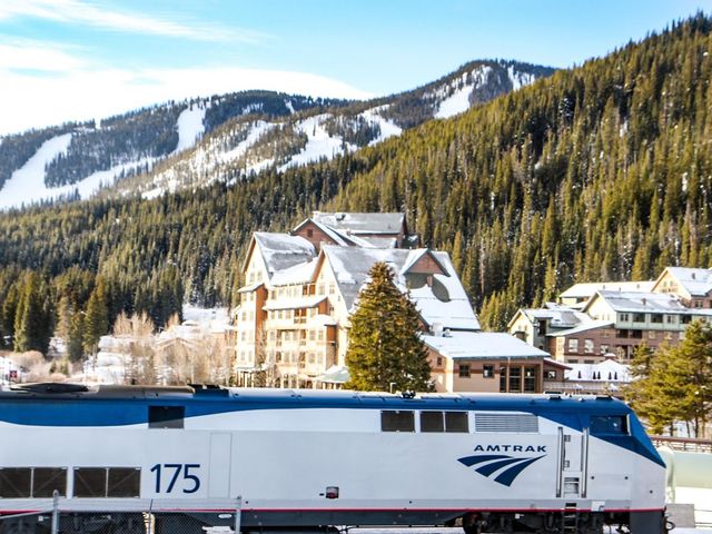 An Amtrak train in front of alpine trees, a snowy mountain, and lodge-style buildings.