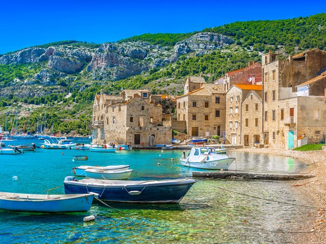 A harbor view in Vis with boats at the shore and old buildings alongside turquoise water and cliffsides dotted with greenery.