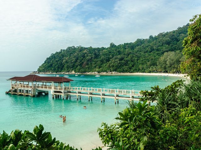 A beach on the Malaysian island of Perhentian Besar with a thick, lush coastline fronting a white sand beach with a pier edging out into the water.