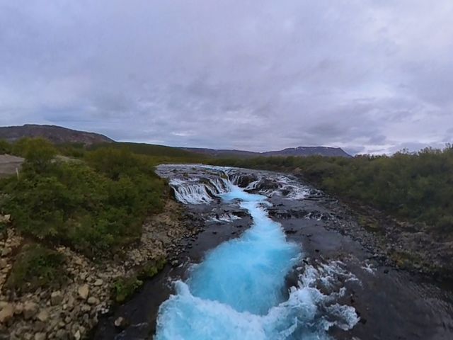A small waterfall feeds into a river with water that glows an electric glacial blue color with rock and brush on either side.