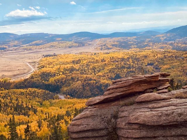 A scenic view over a large landscape as seen from above next to a rock, covered with yellow trees and mountains in the distance.