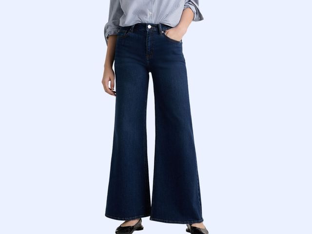 A person is seen from the waist down wearing a button-down shirt and wide-leg blue jeans, against a blue background