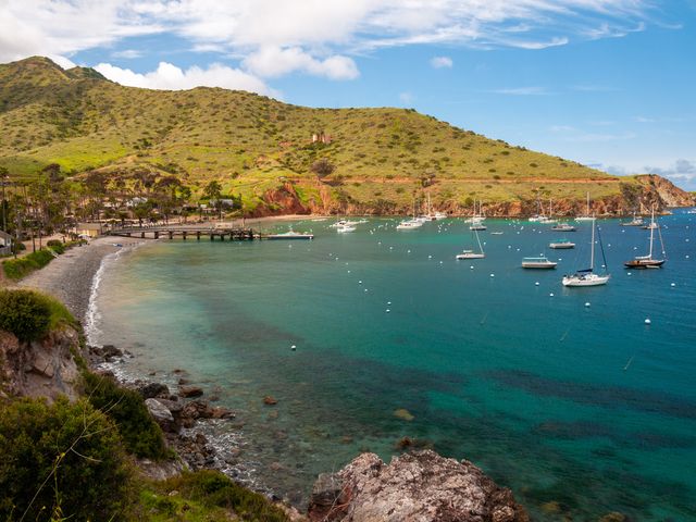 The port of Two Harbors in Catalina Island with a dock, bobbing boats, and a pretty shore area with turquoise waters and green hillsides.