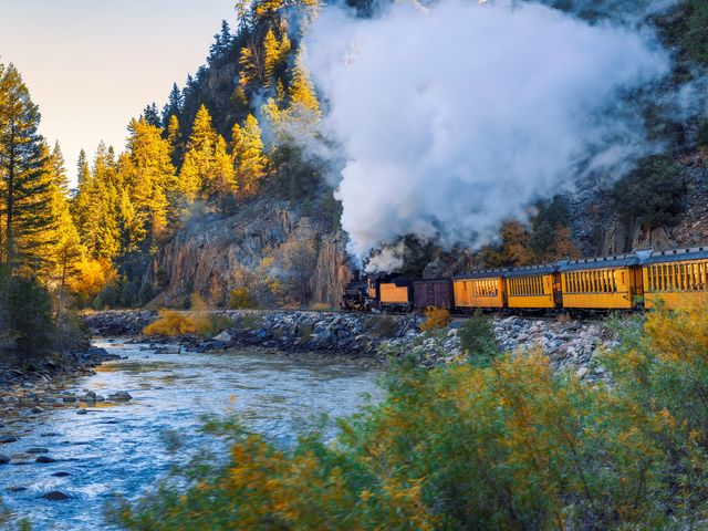 A steam engine train with steam billowing out on a track next to a river with fall foliage.