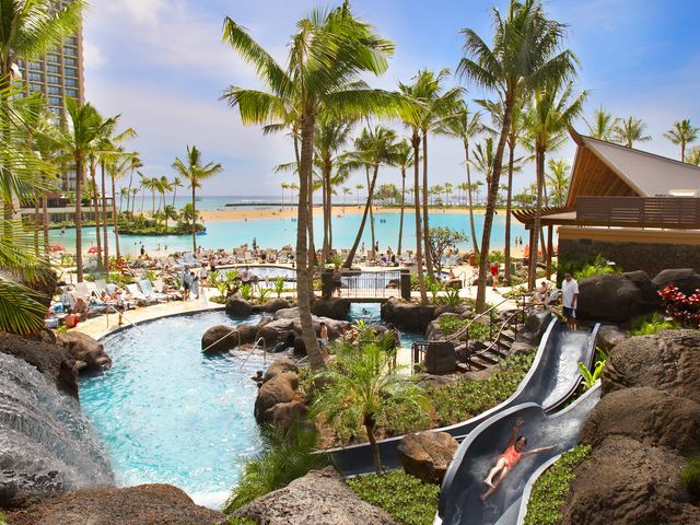 A pool area with someone going down a waterslide framed by palm trees and the ocean in the distance behind it.