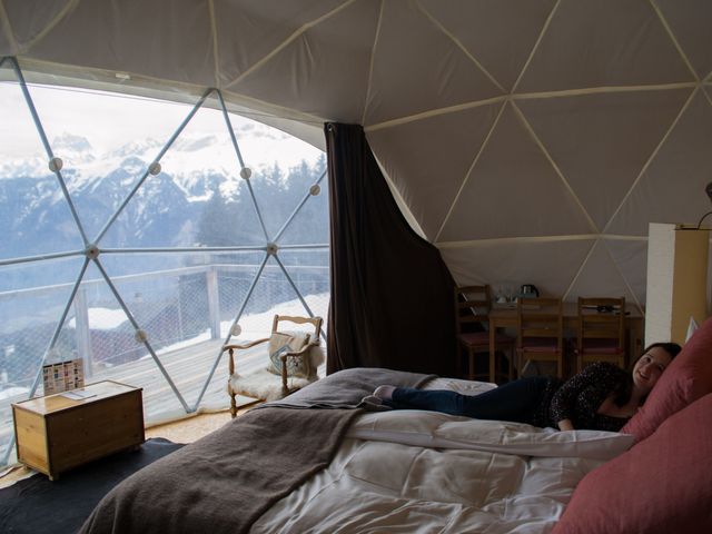 A woman smiles for the camera in bed inside a Whitepod glamping tent in Switzerland with views outside of the snowy Alps.