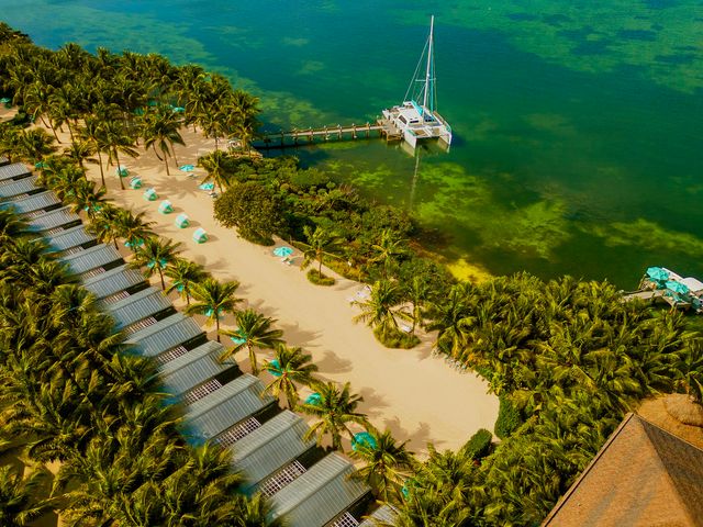 A row of cabins facing the sand on a tropical, lush beach with a dock where a catamaran is parked.