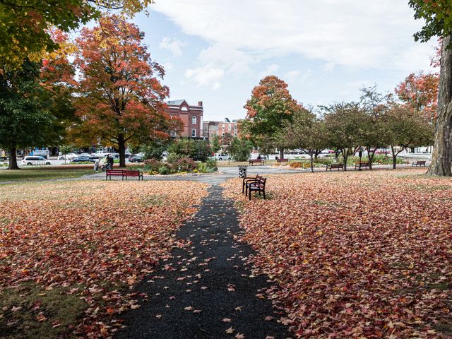 A park in St. Albans, Vermont in fall with changing leaf colors and dried leaves on the ground next to a pathway and park benches.