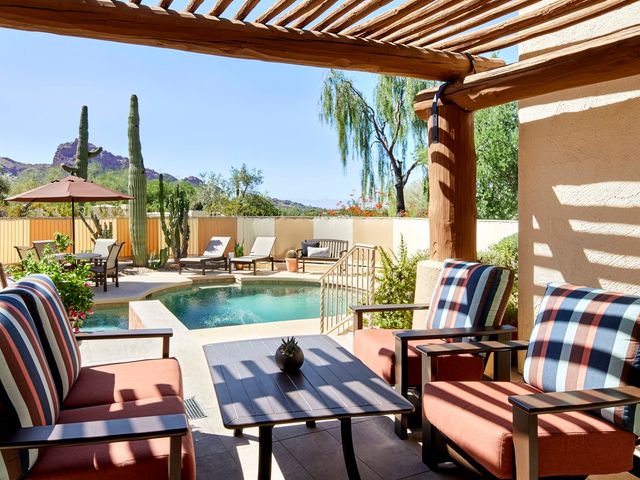 A hotel patio with seating partially shaded by a pergola awning and a small pool surrounded by lounge chairs, cacti, and desert scenery.