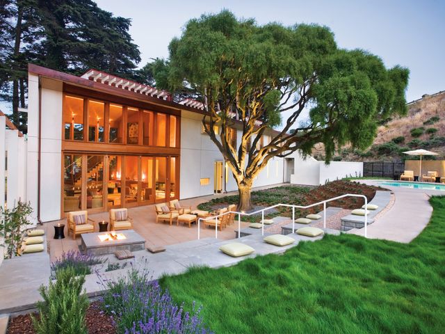 A Mediterranean-style building with large windows facing a nice outdoor terrace, green grass, a big tree, and pool.