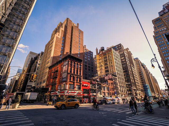 An intersection in Hell's Kitchen in New York City with tall buildings, a taxi, and people walking.