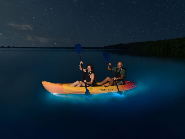 A man and a woman kayak in a double kayak over water that glows blue under a dark starry sky.