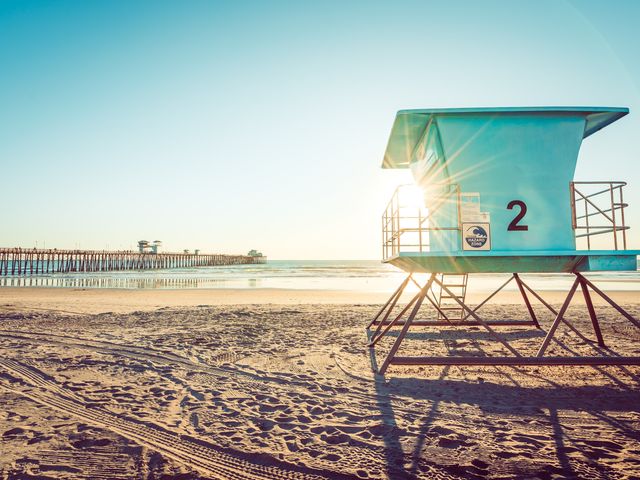 A blue-green lifeguard stand partially obstructs the view of the sun next to a long wooden pier on a wide beach in Oceanside with the tide far out.