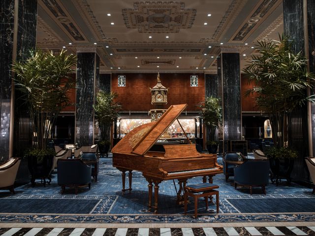 A grand hotel lobby bar with opulent marble columns, an intricate carpet, a long sleek bar in back, and a piano in the middle of the lounge.