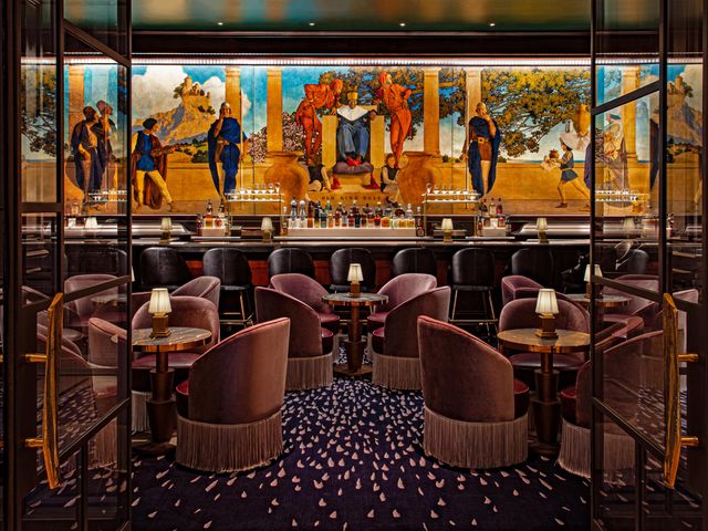 A hotel bar with small round tables, purple velvet chairs, and a large painted mural above the bar.