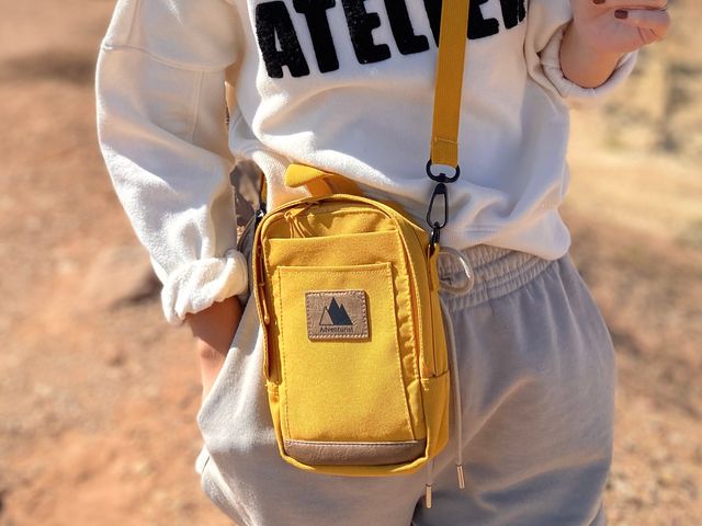 Someone visible only around the waist wearing a yellow crossbody bag with a label that has mountain icons and says 'Adventurist.'