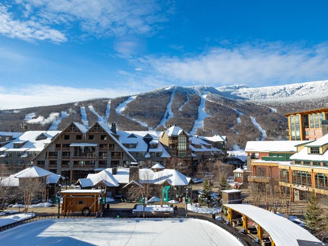 A snow covered resort sits at the base of a mountain with many ski runs, next to a small bustling village.