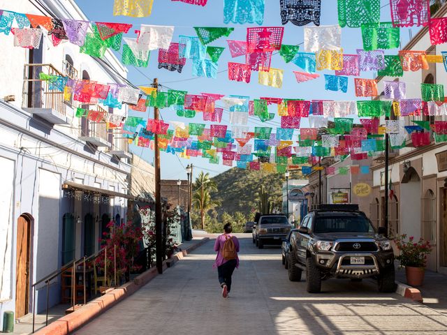 Colorful flags hang over a quiet street with a green hill and palm trees in the background.