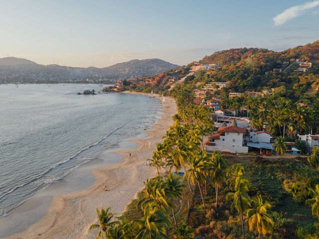 A coastal beach town built into a hillside with red roof buildings and palm trees next to the ocean.