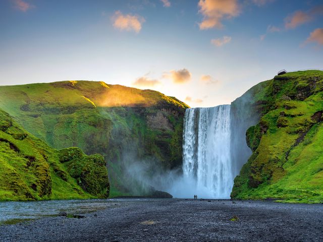 Two people are seen at the base of the enormous Skogafoss waterfall in Iceland with green, mossy mountains on either side of the falls.