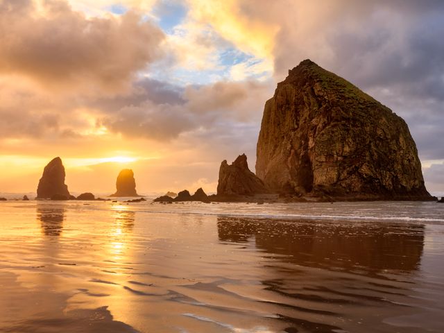 The sun sets behind Haystack Rock on Cannon Beach in Oregon with golden clouds in the sky.