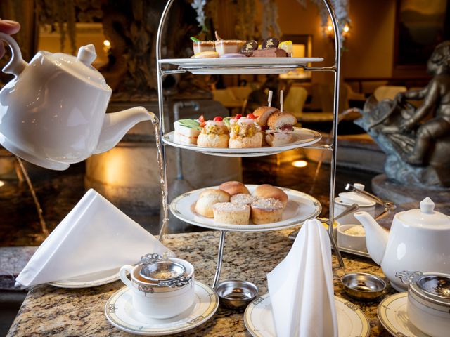 A hand pouring tea from a pot into cups with tiered stands holding small cakes.