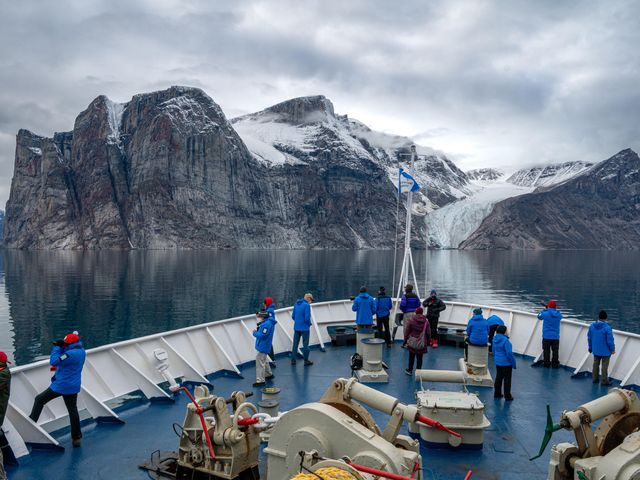 People standing dressed for the cold at the bow of a ship approaching a snowy mountain.