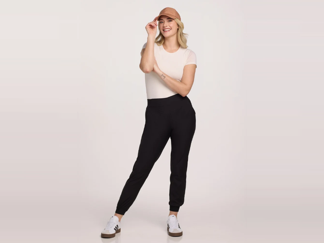 A woman wearing black joggers and a baseball hat against a neutral background. 