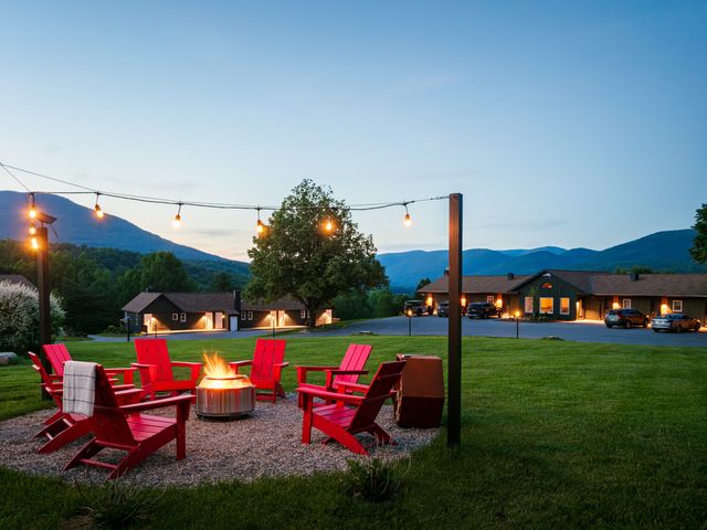 An outdoor firepit encircled by red Adirondack chairs lit by string lights on a grassy area with mountains in the distance.