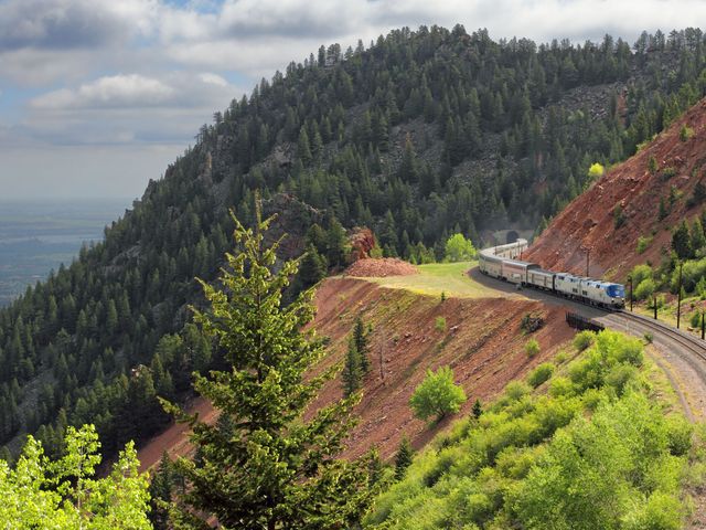A train is seen on the edge of a mountain track in Colorado surrounded by mountains covered with pine trees.