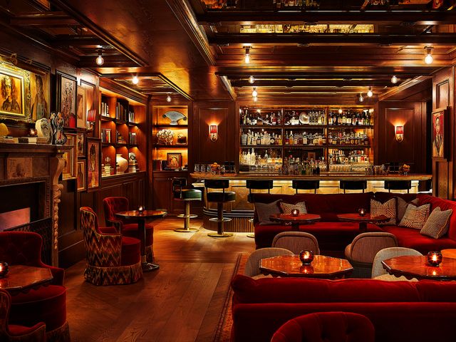 A regal looking hotel bar with dark wood paneling, red velvet couches, a fireplace, bookshelves, and a full bar lit up with sleek lighting.