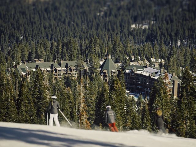 Two skiers on a mountain with views of a large resort next to forested trees.