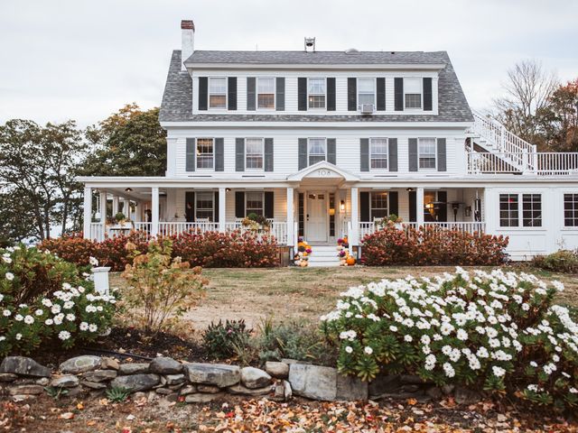 A classic white Colonial home with a front porch and a yard with flowers, surrounded by trees starting to change colors and some dried orange leaves on the ground.
