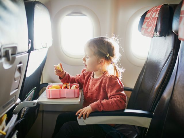 A child sitting in an airplane window seat eating from a snack box.